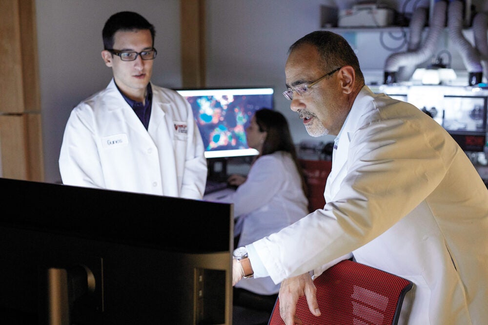 Gökhan Hotamışlıgil leans and points over a computer and points to the screen. Male lab worker in lab coat stands to his left and looks on.