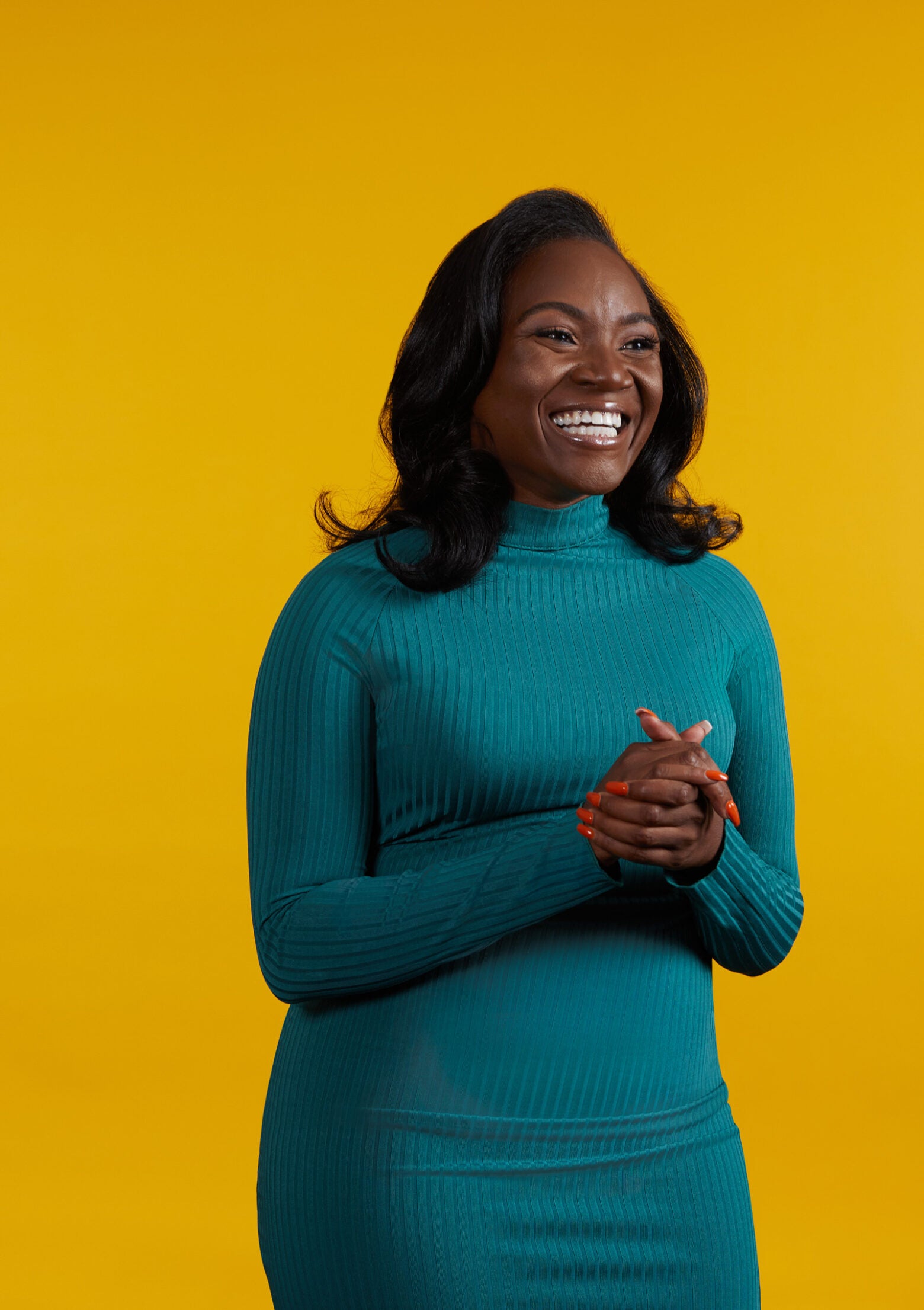 Kizzmekia Corbett wearing a longsleeve, teal turtleneck dress, smiles and looks off camera with her hands clasped.