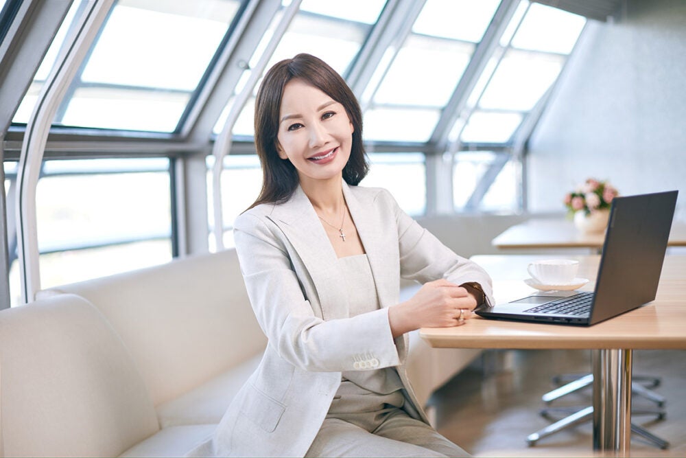 Jane Jie Sun seated at a row of white bench seating, wears a white pant suit with an open laptop beside her, smiling at the camera with her hands rested on the table. The room has large windows and natural light shines around her.