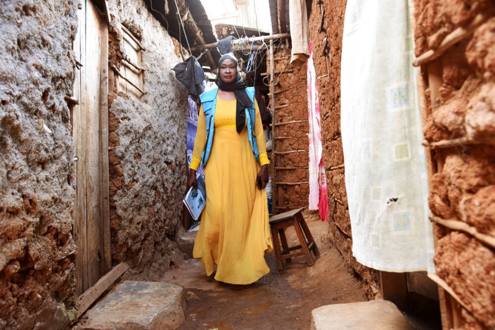 A tall woman wearing a yellow dress and blue scarf/sari walks down a dirt alley between two stone buildings in Nairobi's Kibera district. She holds a cell phone and notebook..