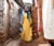 A tall woman wearing a yellow dress and blue scarf/sari walks down a dirt alley between two stone buildings in Nairobi's Kibera district. She holds a cell phone and notebook..