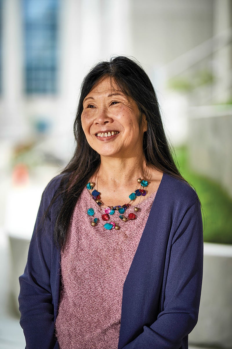 Professor Phyllis Kanki stands outside on a concrete walkway with greenery behind her. She wears a purple cardigan, light pink shirt and colorful beaded necklace. She has black hair that extends past her shoulders and a natural smile.