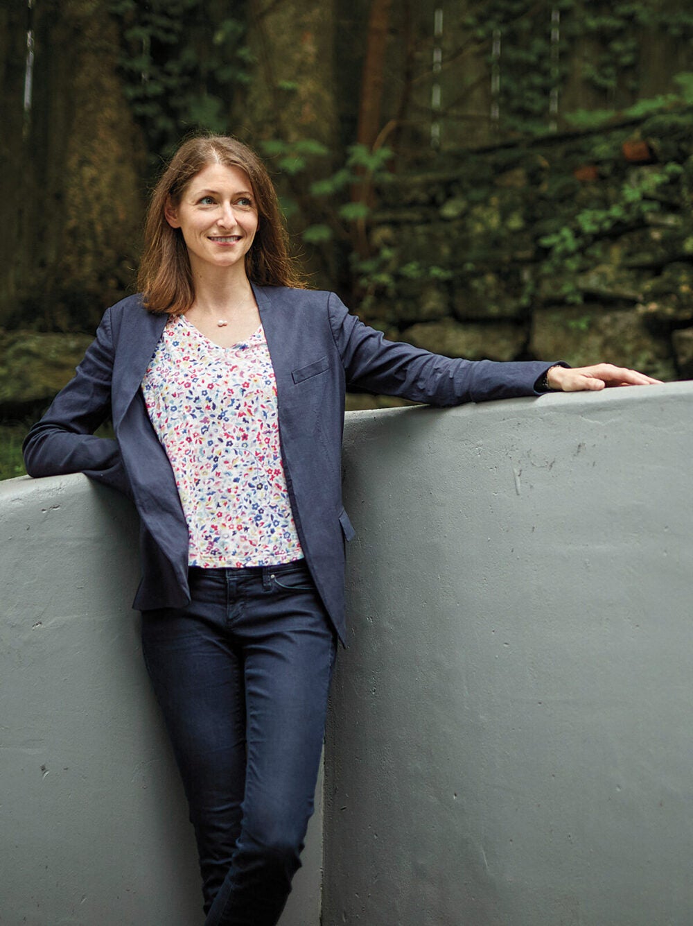 Nora Kory stands in a wooded area by a short concrete wall, resting one elbow on one side, and extending her arm on the other. She wears jeans, a multicolored shirt and a dark blue blazer.