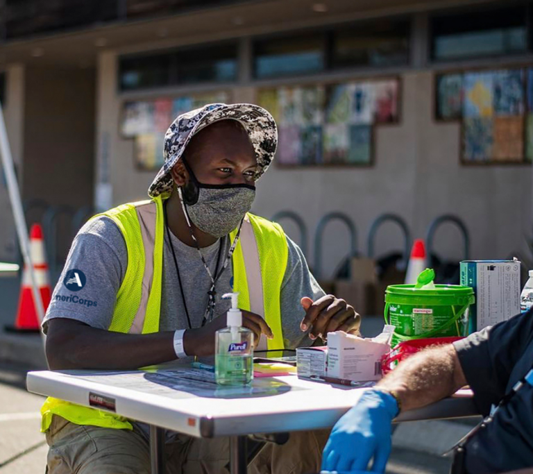 A black man wearing a sunhat, facemask, yellow caution vest and grey AmeriCorps t-shirt, sits outside at a white table with PPE and handsanitzer.