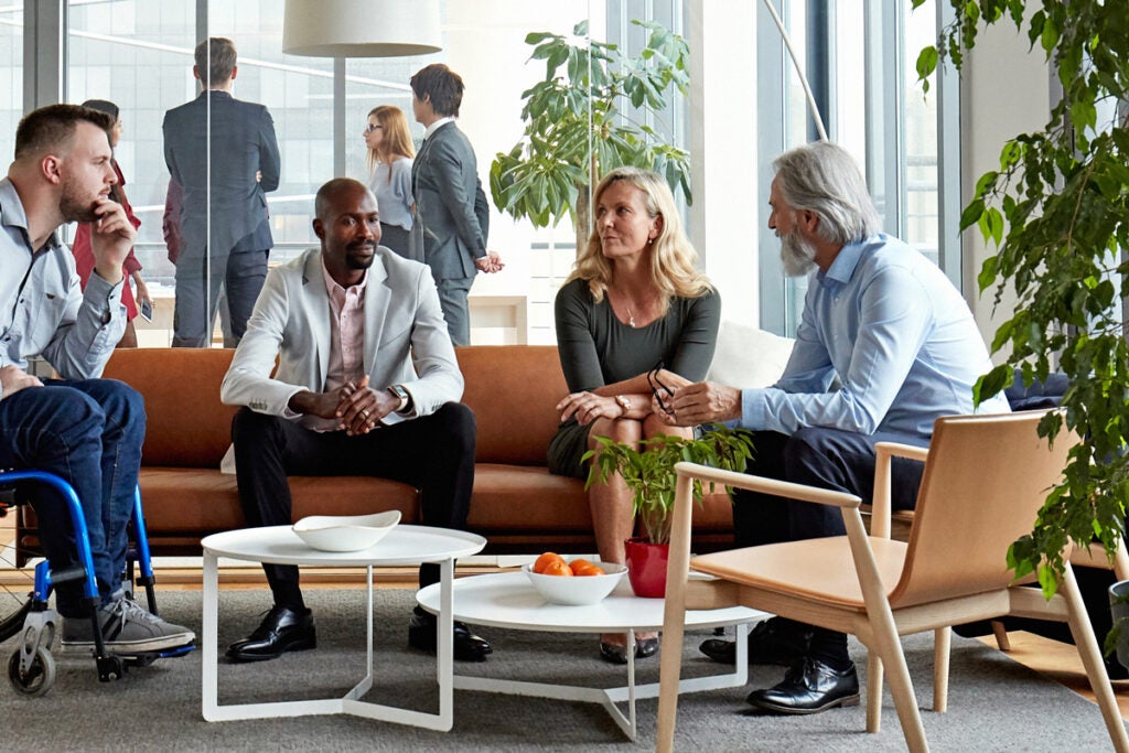 Four employees sit in a lobby area in an office. They are dressed in business casual clothes, sit on an orange couch, and arm chairs. There are a couple large plants in the area.