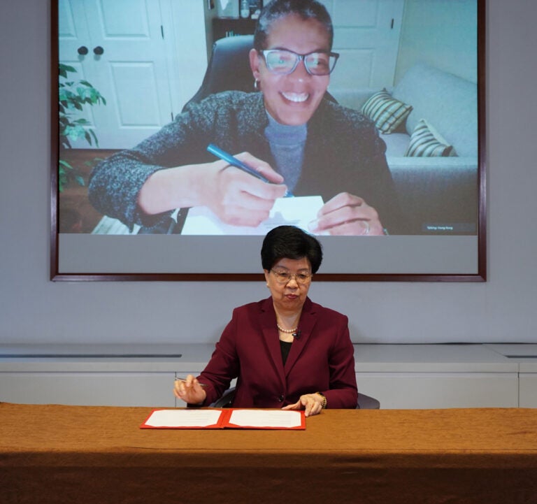 Michelle Williams, projected on a large screen, holds a pen, ready to sign an document. In the foreground sits Margaret Chan, dean of Tsinghua Vanke School. She sits at a large brown desk and has a document portfolio and also holds a pen.