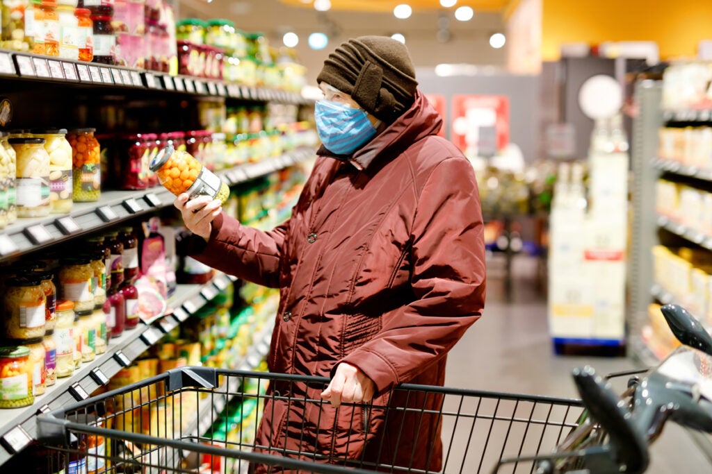 A older human stands in a grocery aisle with a cart, holding and looking at a can of preserves. The person wears a crimson winter jacket, brown knitted hat and a face mask.