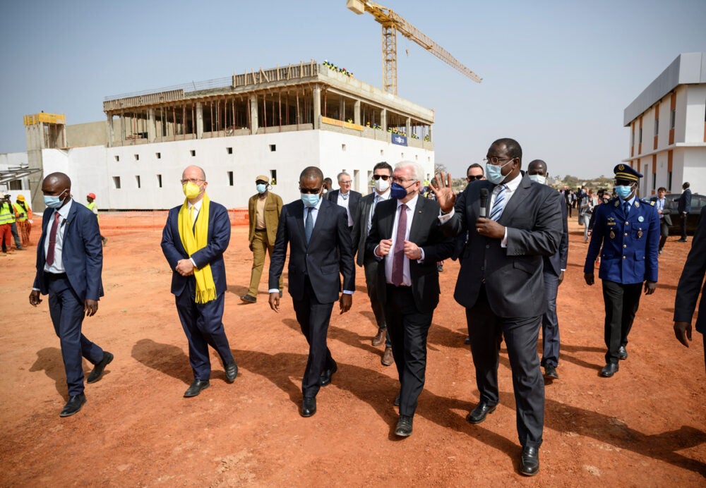 Four governmental officals wearing suits and face masks walk on orange dirt, conversing. A building under construction with a crane and white tacking is behind them. Other government officials and men in uniform walk behind.