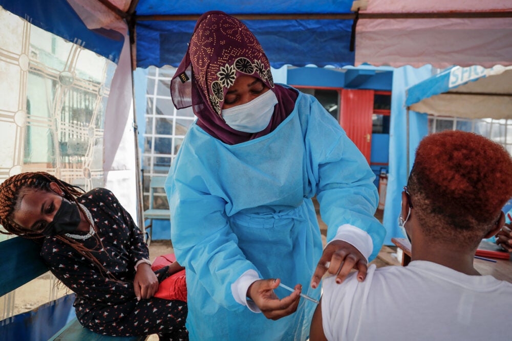 In an open-air blue tent, an African woman wearing a blue surgical gown and maroon printed head scarf gives a shot to a seated person wearing a white shirt with close cropped har. A young woman with braids, a maroon pattered dress and a mask looks on.