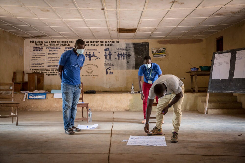 Three african men, wearing masks, stand is an open room in a school with wooden chairs behind them. They are looking at a large white piece of paper on the floor with writing. Other pieces of large paper are in the background with writing.