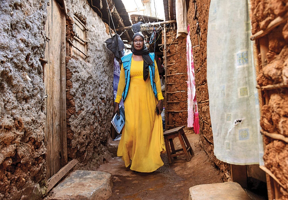 A tall woman wearing a yellow dress and blue scarf/sari walks down a dirt alley between two stone buildings in Nairobi's Kibera district. She holds a cell phone and notebook..