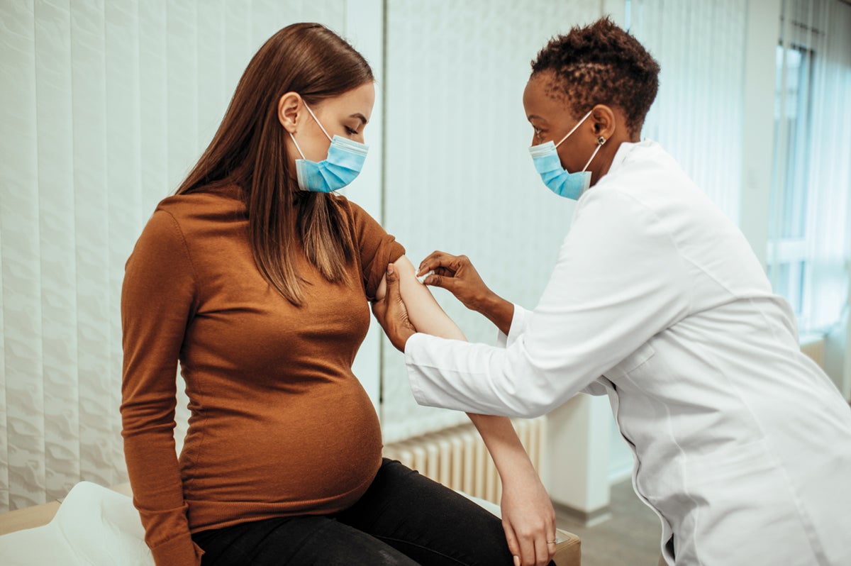 Seated brunette pregnant woman wearing an orange turleneck receiving medical shot from female medical professional wearing a while labcoat.