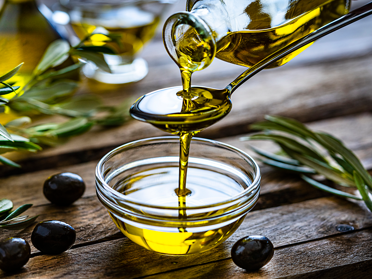 Olive oil is poured from a glass jar into a spoon into a glass bowl. It rests on a wooden table with olive leaves and olive fruit.