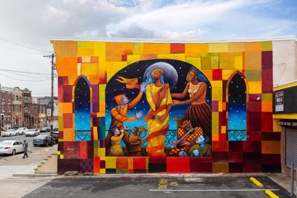 Mural entitled "Resilience". Three dark figures wearing orange and yellow dresses stand in an archway outdoors. The middle figure is in front of the moon and has her head turned 90 degrees. The archway is made up of shades of orang, yellow and grey blocks. The outside implies twilight with stars.