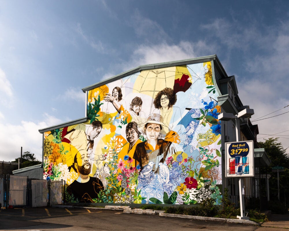 Mural entitled "Brighten the Corners. Located at 114 West Queen Lane." On the side of a set of homes, community figures are painted and intertwined with birds, umbrellas, and flowers. Figures are a mix of male and female and smile or gaze at the viewer.