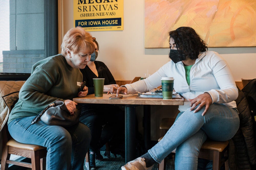 Megan Srinivas sits at a table wearing blue jeans, a white zippered jacket, green shirt and facemask, and talks with two volunteers over a cell phone. Cups of to-go coffees sit on the table.