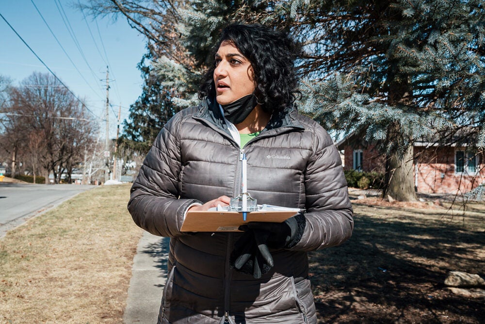 Megan Srinivas— black hair, brown skin and wearing a grey parka—holds a clip board and cellphone while walking in a neighborhood