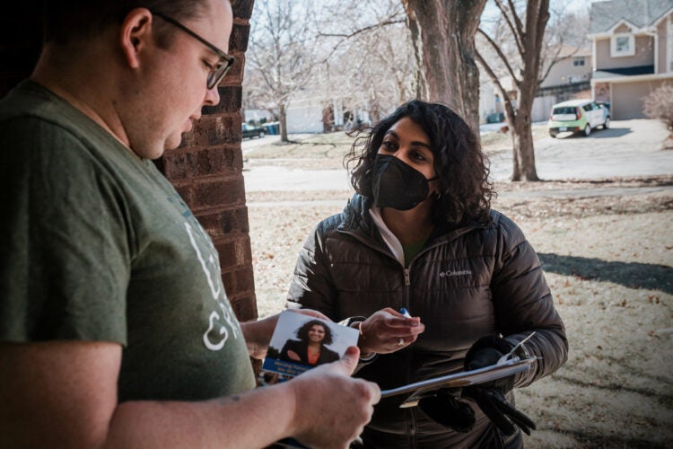 Megan Srinivas— black hair, brown skin and wearing a grey park and black face masks—talks with a constituenton his front stoop in a Iowa neighborhood.