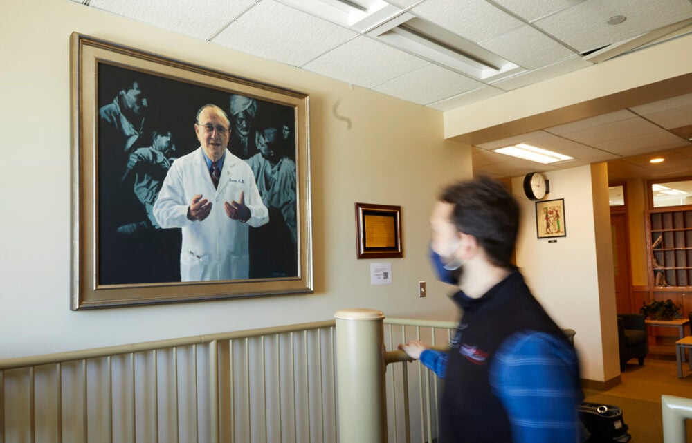 A male student wearing a blue shirt, black vest and black facemask walks past a portrait of Bernard Lown.