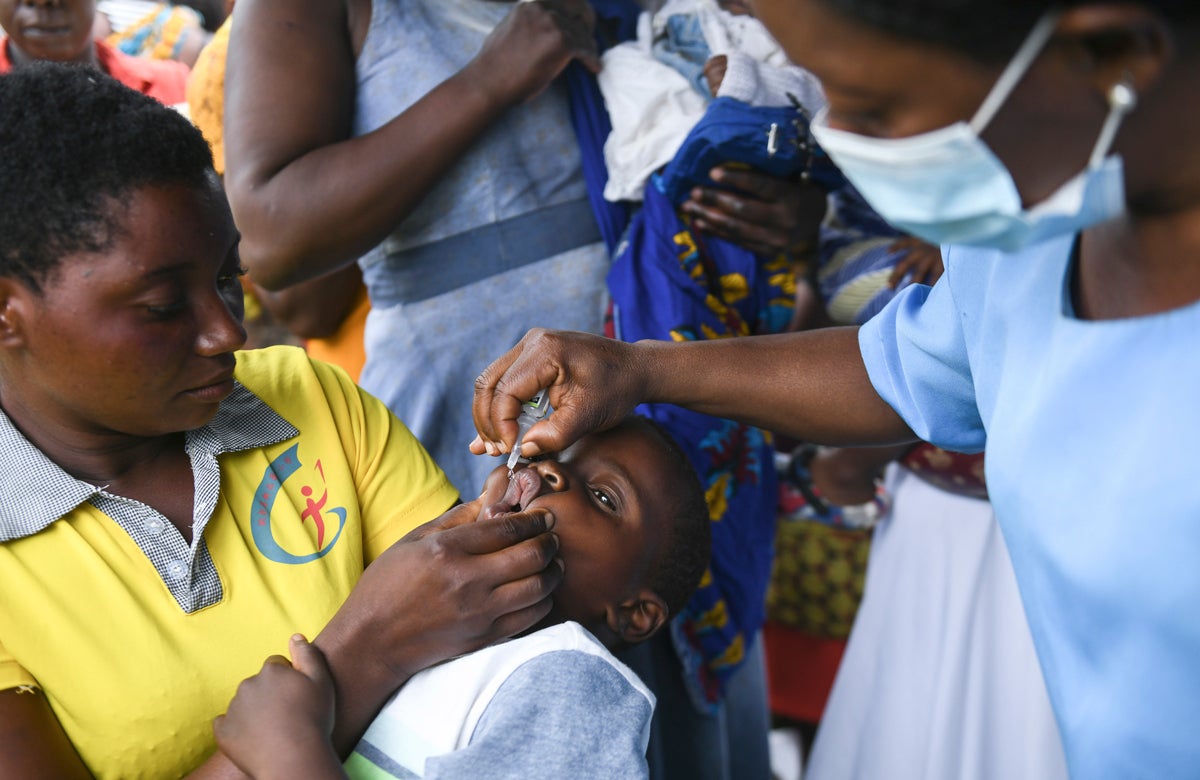 A child recieves an oral polio vaccines in Malawi. She is in the arms of a woman wearing a yellow shirt, and a medical professional in blue scrubs and facemask administers the test.