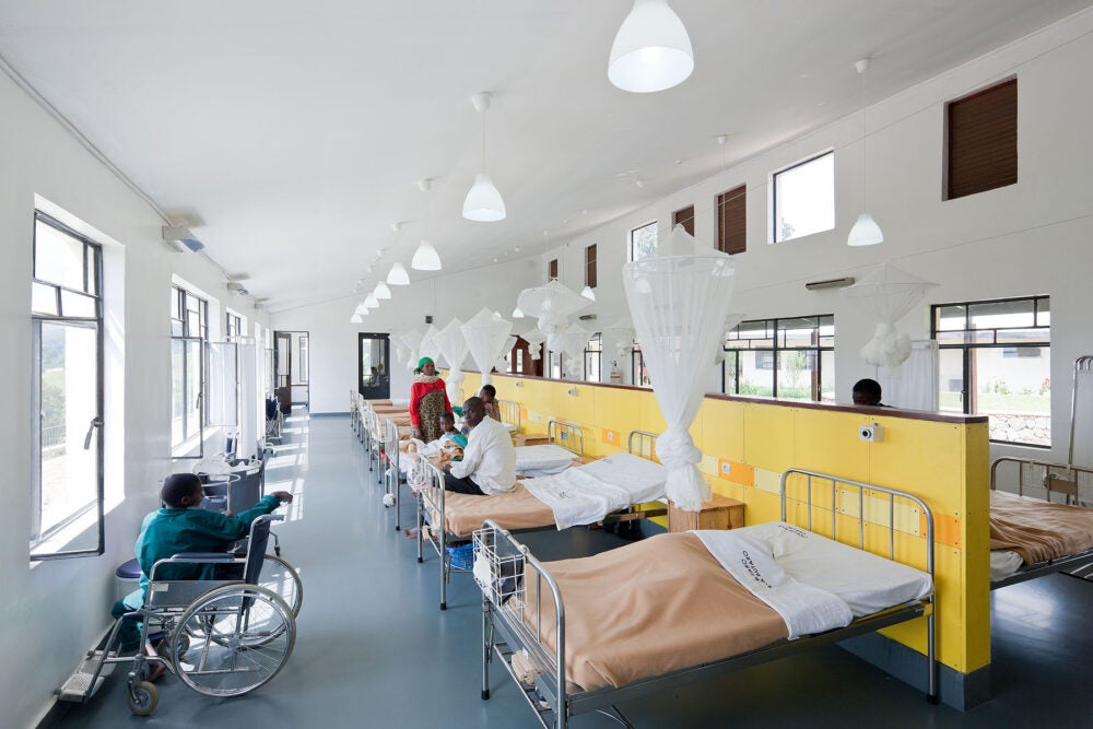 The Butaro District Hospital in Rwanda: a short yellow wall divides a large room with evenly spaced hosptial beds on each side. Mosquito nets hang above each bed. White lights and many windows make the space bright. Patients sit on beds or in wheelchairs along the wall.