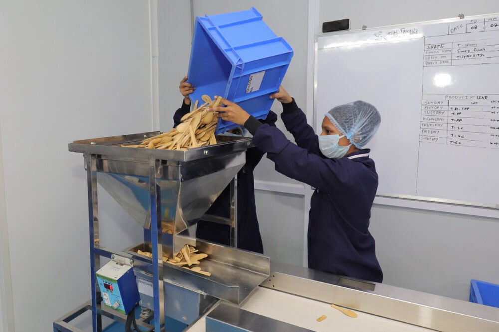 A woman wearing a navy long sleeve shirt, hairnet and facemask, pours edible spoons from a blue bin into a metal funnel along a conveyer belt in a factory