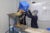 A woman wearing a navy long sleeve shirt, hairnet and facemask, pours edible spoons from a blue bin into a metal funnel along a conveyer belt in a factory