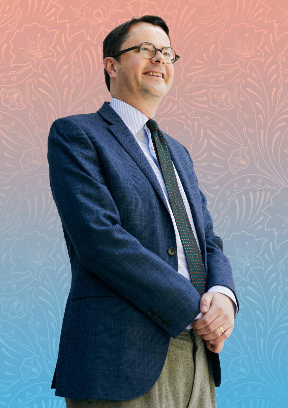 Tyler VanderWeele in khaki pants, a blue blazer, white shirt and tie with his hands clasped and smiling off into the distance. He stands in front of a paisley patterned background, colored from pink to blue.