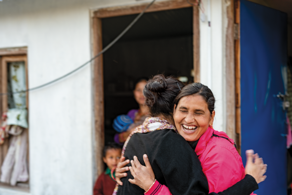 Isha Nirola in a black sweater and white patterned scarf is embraced by a Nepalese woman weraing a pink jacket and a large smile. A Nepalese family looks at them in a doorway.