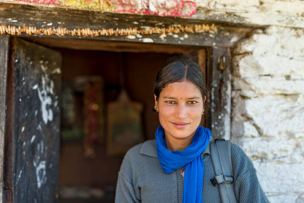 A female Nepalese clinician stands outside a doorway wearing a blue scarf and backpack.