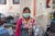 A female Nepalese clinician stands in a hospital room, wearing a facemask, pink cardigan, teal shirt and patterned scarf, holding bags of saline solution.