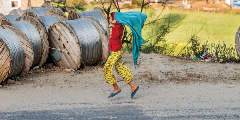 A girl dressed in bright yellow pants, blue sandles, red hoodie runs on a rural gravel road, holding a teal blanket over her head like a cape.