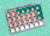 An unopened pack of birth control pills rests on a light teal background. The pills are in four rows of seven; three rows have orange pills, the last row has white pills.