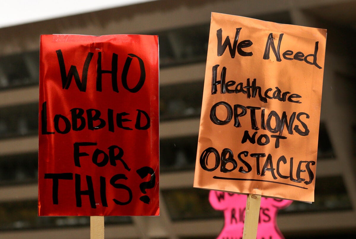 A red protest sign (left) and an orange protest sign (right) are held in the air. The red one reads “Who lobbied for this?” in black text. The orange one reads “We need healthcare options not obstacles.”