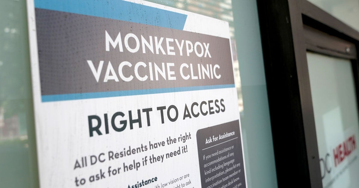 A blue, white and gray sign reads “Monkeypox Vaccine Clinic Right To Access”. It it taped to a storefront window. A “DC Health” logo is affixed to an entrance door in the lower right-hand corner of the image.