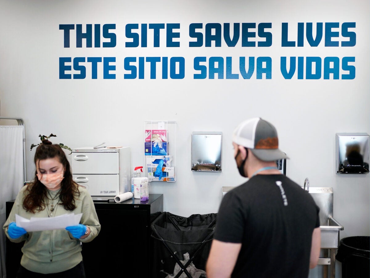 Bold blue text on the wall reads "This site save lives" in Spanish and English at an overdose prevention center. Below the sign are metal paper towel dispensers, sterile blue golves and a copy machine. A young woman with brown hair, organge face mask and blue gloves holds and reads a peice of paper. A man wearing a black t-shirt and backwards baseball hat faces her.