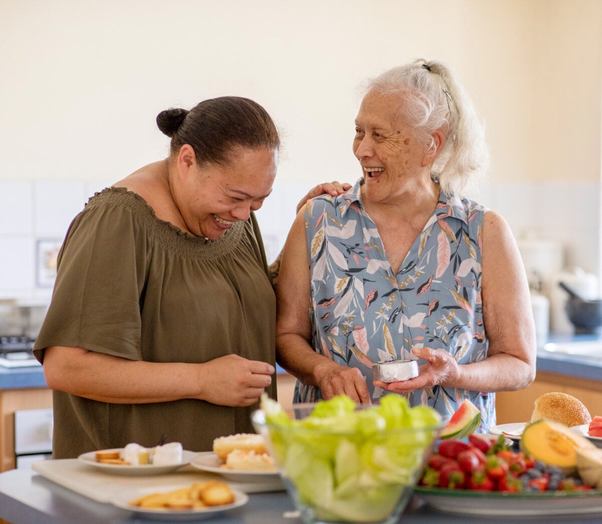 Two older brown women stand at a kitchen island smiling and laughing while preparing food. Fruit and cheese are in the foreground.