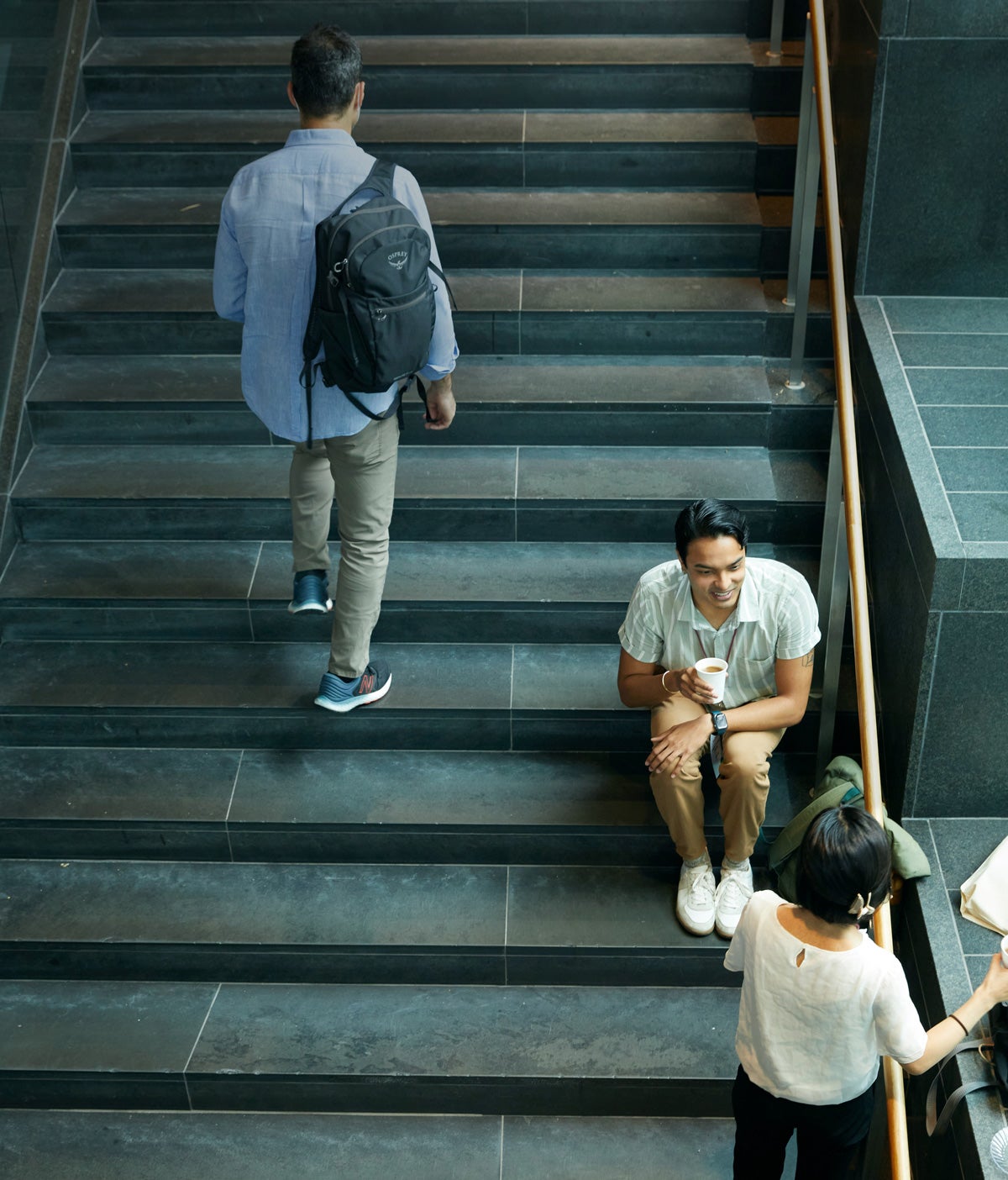 A male student sits on a large dark marble staircase while talking to a female student who stands. A third person in the top right corner of the photo ascends the stairs.