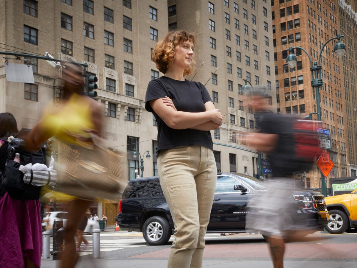 Cristina Gago stands outside in New York City, wearing a black t-shirt and tan pants. Her arms are folded and she's looking to her right. Cars and people are blurred in the background.