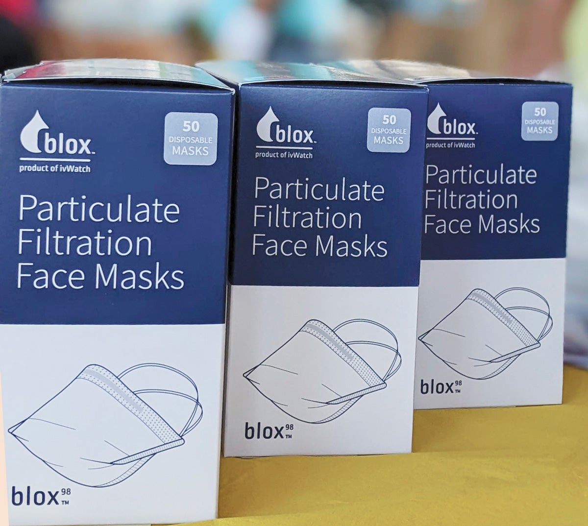 Three boxes blue and white boxes of Particulate Filtration Face Masks sit on a yellow table.