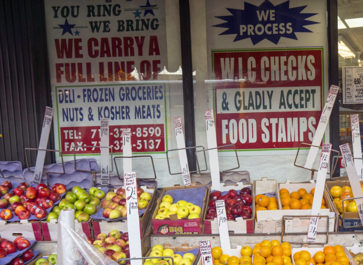 A bodega window displays a sign that reads: We process W.I.C checks & gladly accept food stands.” Various fruit are in fruit stands boxes in front of the window.