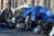 Blue tarps, tents, bikes, strollers and other items are strown on a sidewalk forming a homeless camp in an area commonly known as Mass and Cass in Boston. A feamle wearing a black sweater and holding a cigarette looks into tent.