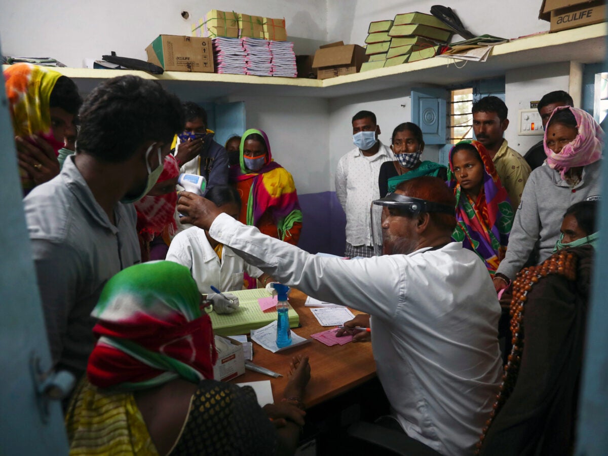 Mannanur in Nagarkurnool District of Telangana State, India: A doctor sits at a desk in a small, crowded exam room at a medical clinic. The doctor has a clear face shield and is extending a digital forehead to check a person’s temperature. The people are wearing bright patterned scarves and head wraps. Most wear face masks.