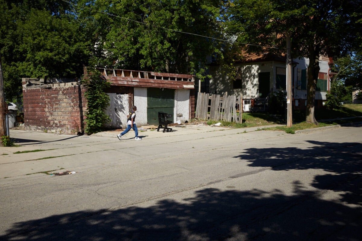 A Black child walks along a street in the Metcalfe Park neighborhood in Milwaukee. He’s passing by a rusted garage, with paint peeling on one side, ivy growing on one corner. An abandoned black shopping cart is by the garage. A wood fence falls apart on one side. Trees are overgrown.