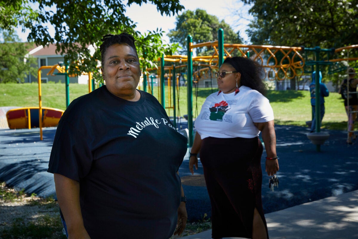 Danelle Cross and her adult daughter Melody McCurtis stand in front of a playground at a green park. Cross, at the left, looks directly at the camera. She is Black and wears a black t-shirt with the words “Metcalfe Park” on it. McCurtis, right, is in the background, looking off camera, and wears a white-tshirt and a long black skirt.