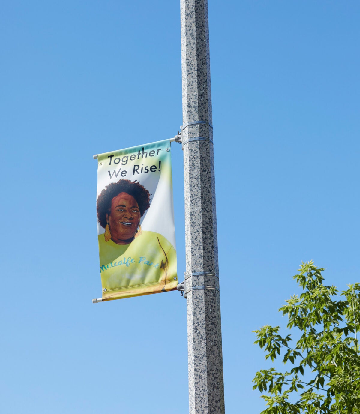 A street post hoists a white and green banner with an illustration of Danelle Cross and the words “Together We Rise.”