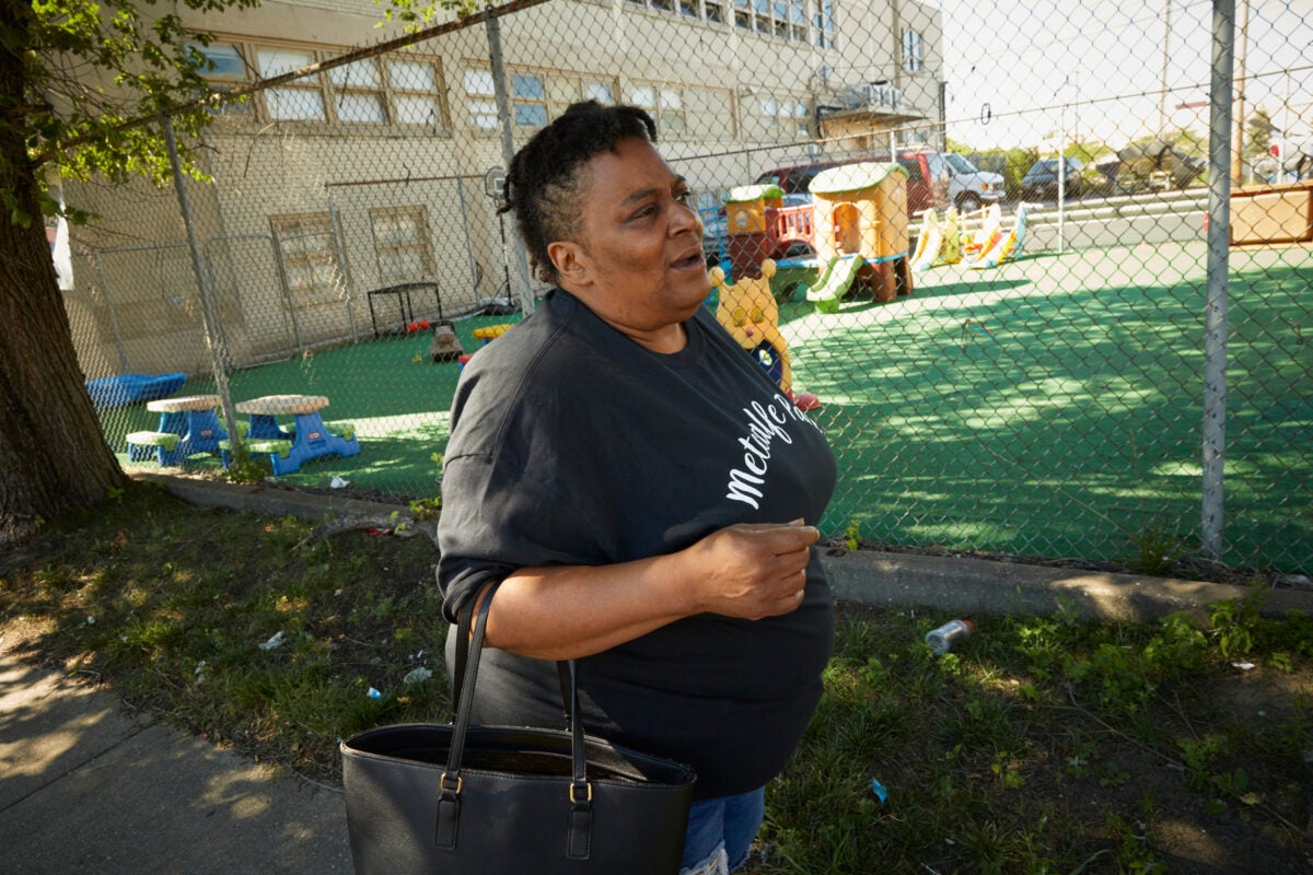 Danelle Cross walks along a street in the Metcalfe Park neighborhood in Milwaukee, Wisconsin. A daycare greenspace is behind her. She is Black, wears a black “Metcalfe Park” T-shirt , and carries a large black handbag over her arm.