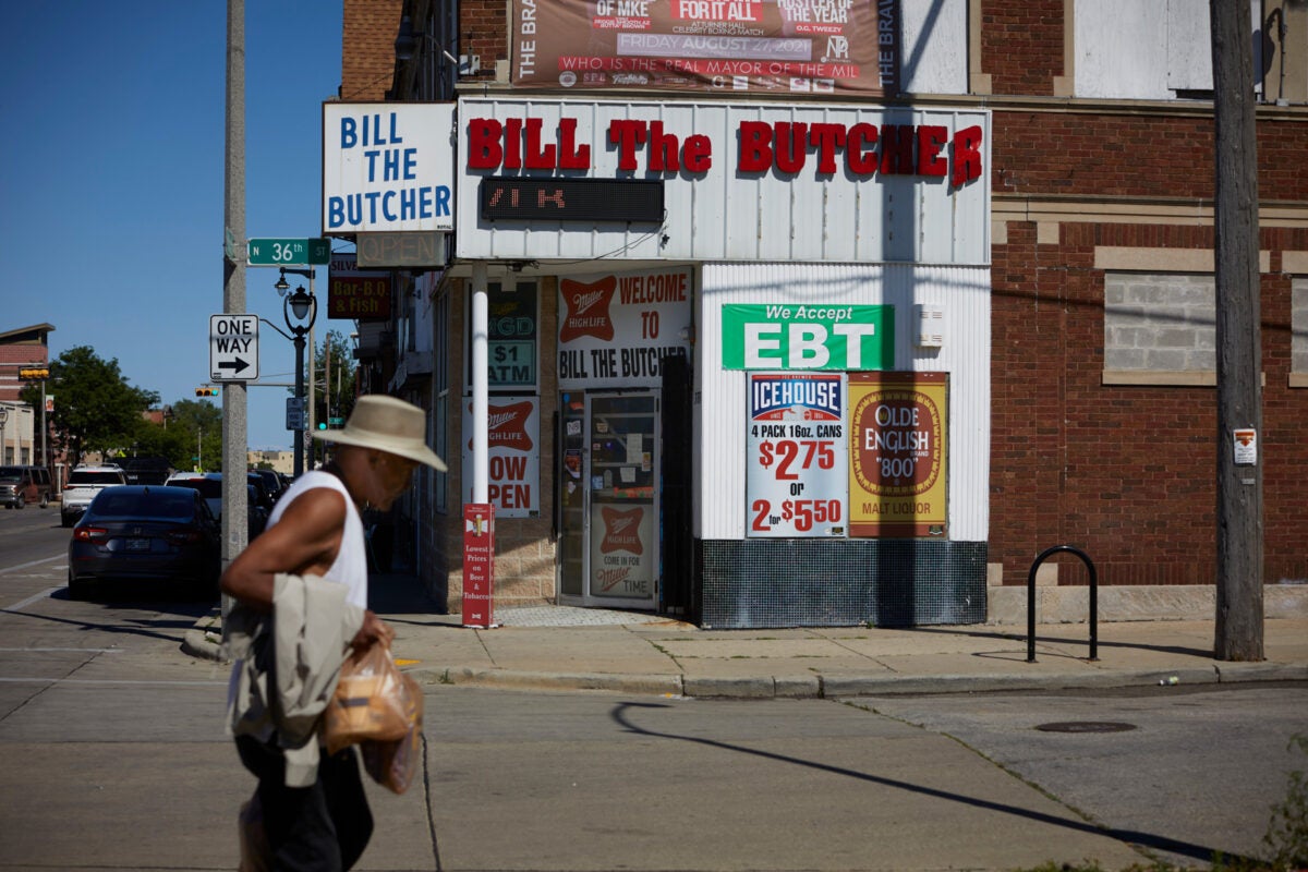 An exterior photo of a corner story in Milwaukee’s Metcalfe park neighborhood. “Bill the butcher” is in large red letters on top of white paneling, and a white corner marquee also reads “bill the butcher” in blue writing. A green EBT sign, and beer and cigarette advertisements are paneled on the building. A man wearing a t-shirt, straw sunhat, holds a bag, and walks with a cane in the foreground.