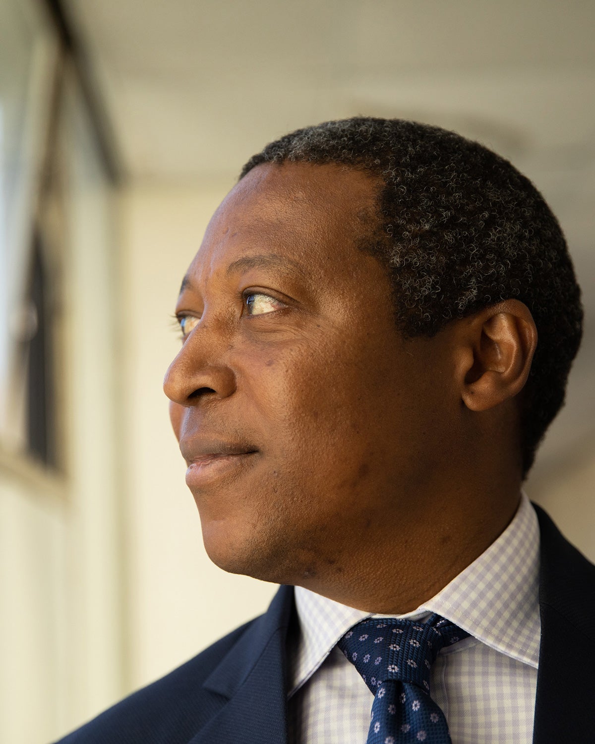 Ntobeko Ntusi looks out a window in a non-descript buliding. Windows are open behind his shoulders showing a blue sky. He wears a navy blue suit, white shirt and tie. He is black and has close-cut hair.