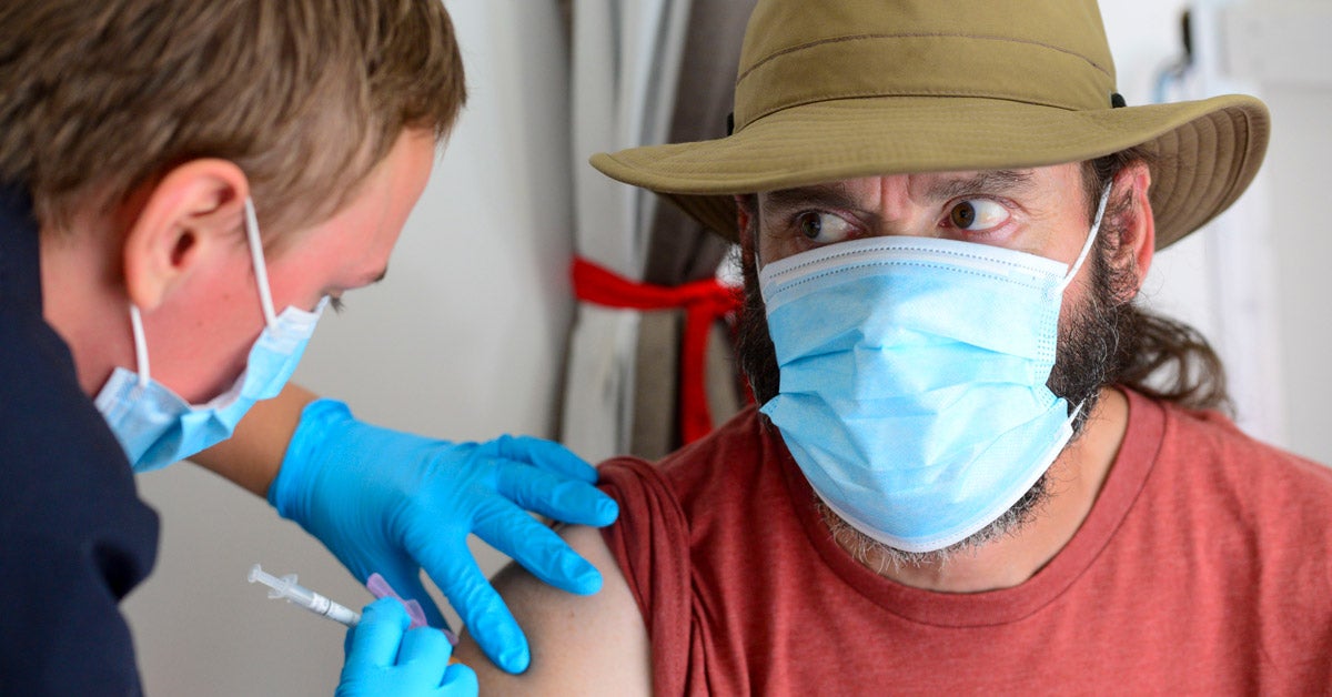 A middle-aged white man, wearing a wide-brim tan hat, red shirt, and blue facemasks, gets a COVID-19 shot by a technician wearing blue latex gloves and a facemask.